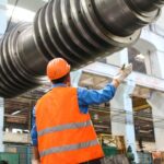 A factory worker in safety gear gestures towards large industrial machinery, representing the robust work ethic in Europe's manufacturing sector.