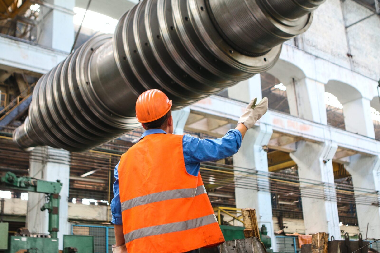 A factory worker in safety gear gestures towards large industrial machinery, representing the robust work ethic in Europe's manufacturing sector.